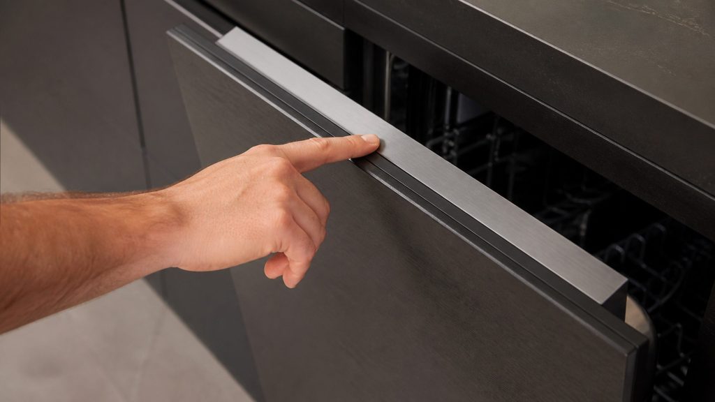A finger pressing a blank touch control on the top edge of a fully integrated dishwasher door, the door ajar by an inch