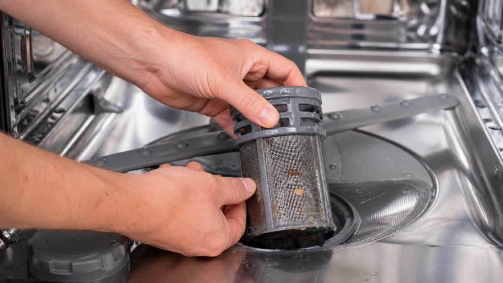 Two hands lifting the cylindrical mesh filter out of the floor of a dishwasher tub, food debris caught in the mesh