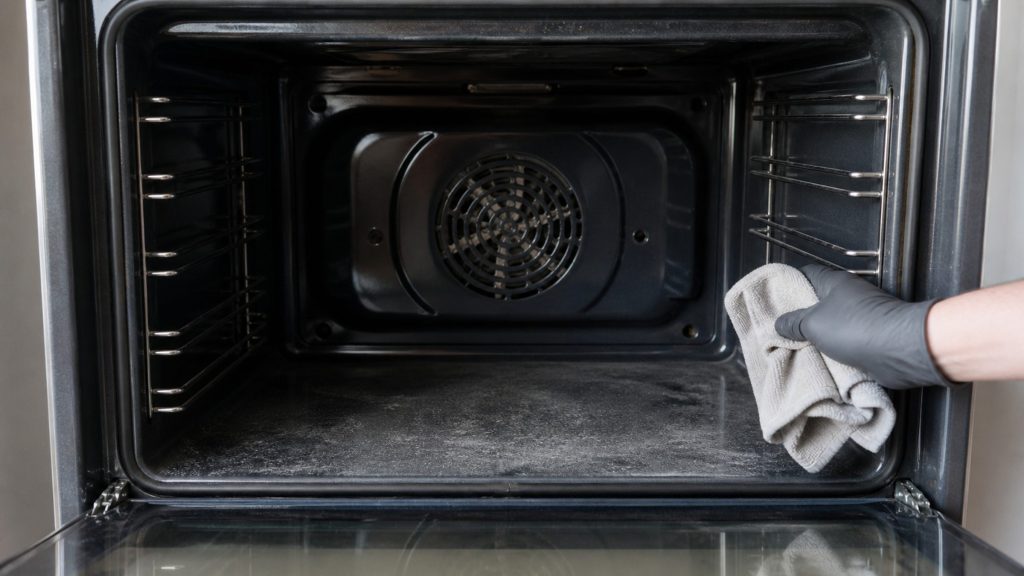 The empty cavity of a built-in oven with the door down, fine gray ash on its floor and a gloved hand wiping it with a cloth