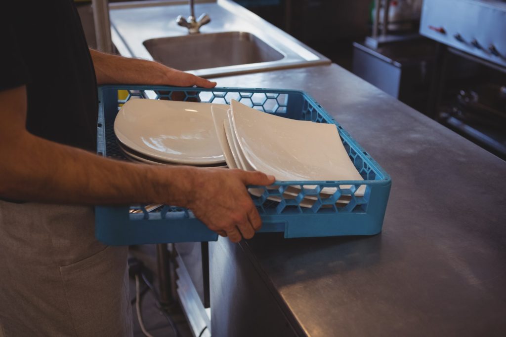 Two hands carrying a blue dish rack of stacked plates along a stainless bench