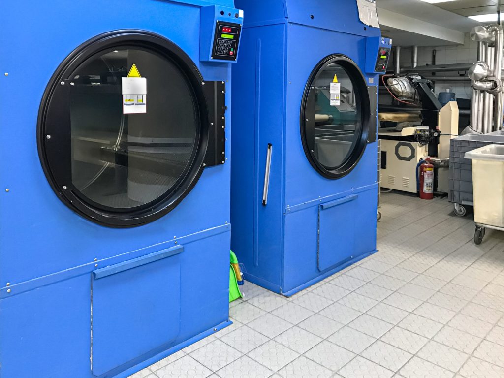 Two large industrial tumble dryers with round glass doors in a working laundry, an ironer behind them
