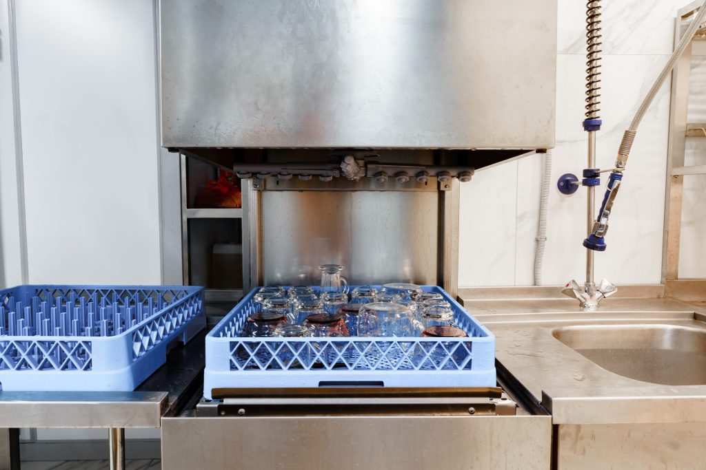 A hood-type dishwasher with its hood raised over a rack of glasses, a second rack waiting at the left