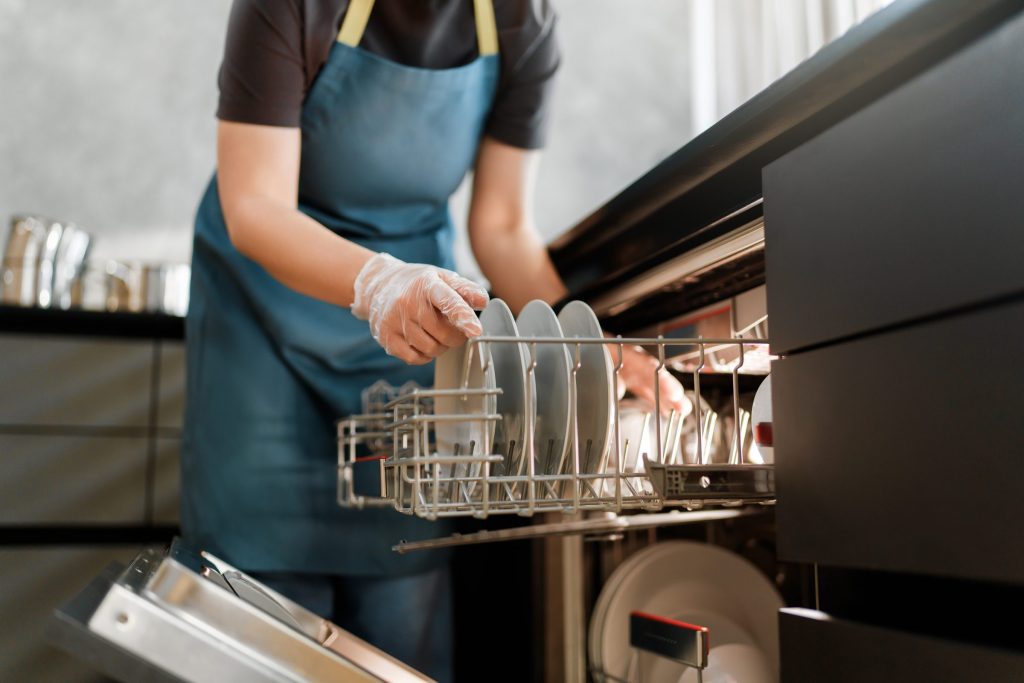 A kitchen worker in an apron loading plates into the rack of a built-under dishwasher behind a dark front