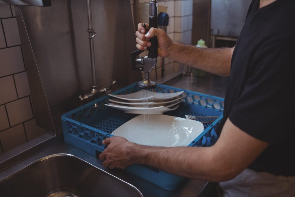 Plates in a blue rack under a pre-rinse spray arm at a stainless sink