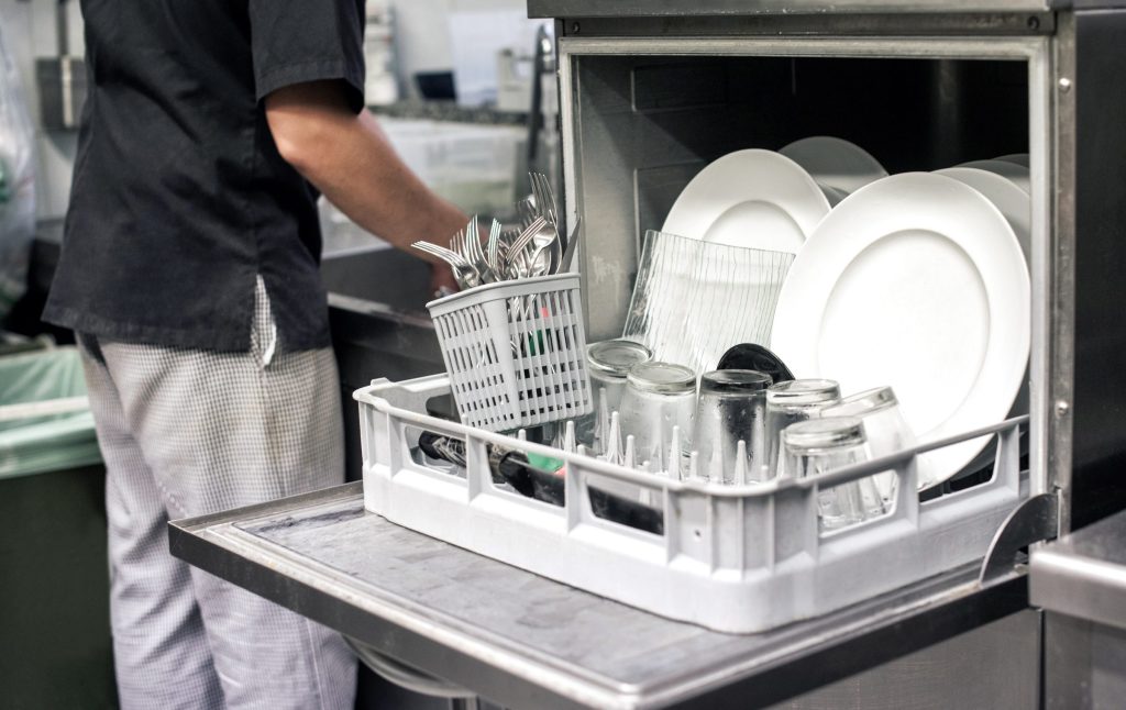 A kitchen porter sliding a loaded rack of plates and glasses into a stainless commercial dishwasher