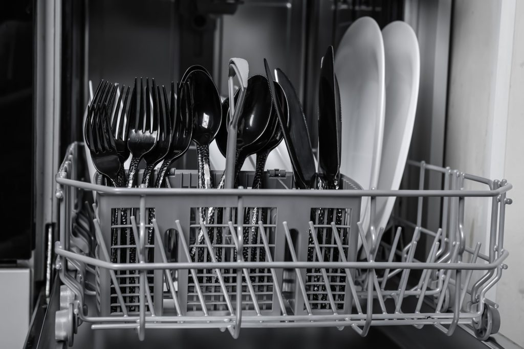 The cutlery basket of an open dishwasher, forks and spoons upright, plates behind