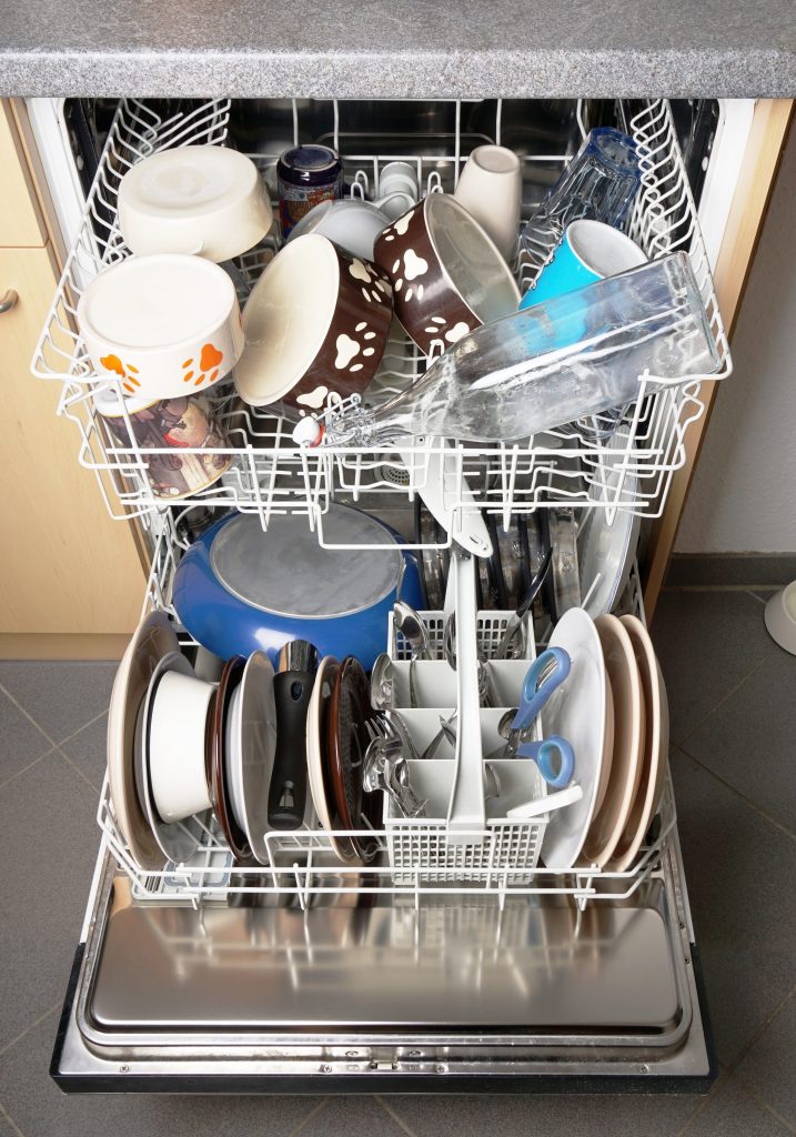 A dishwasher door down with both racks loaded, plates on edge below and cups above
