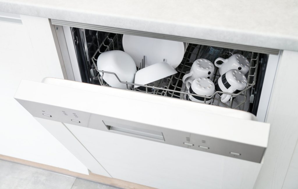 A panel-ready dishwasher ajar behind a white cabinet front, white cups and bowls in the upper rack