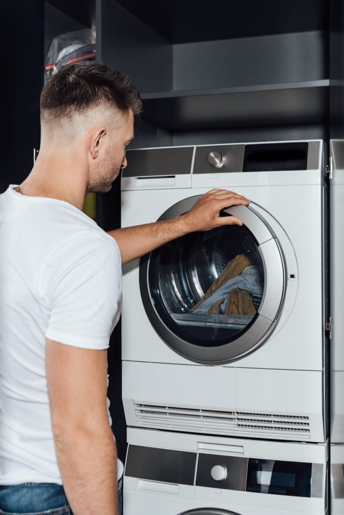 A man at a stacked laundry pair in a dark cabinet, the washer door open on a load