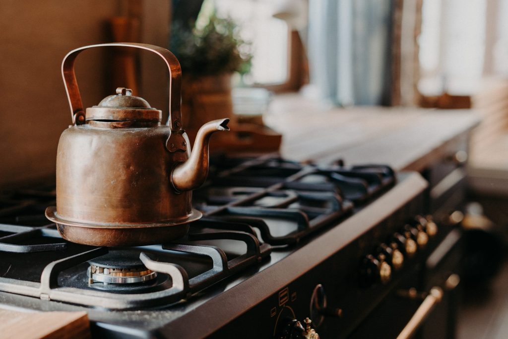 A copper kettle standing on the cast grate of a dark professional range with brass knobs