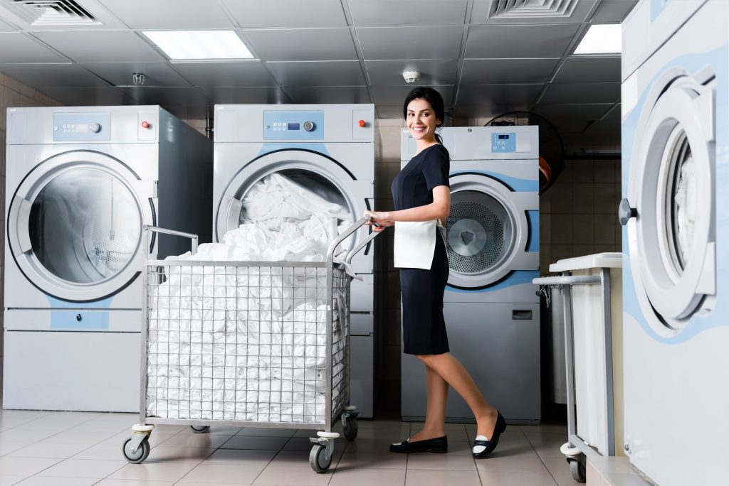 An on-premise laundry: white professional washers and dryers along a tiled room, a linen cart of white sheets being wheeled past