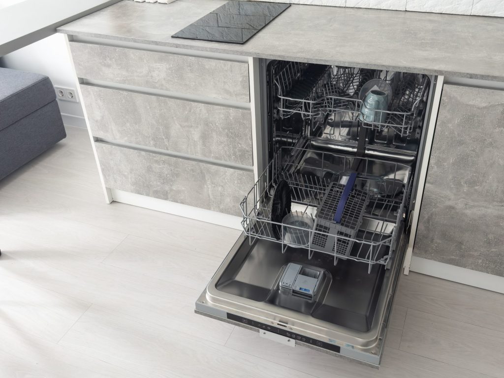 A fully integrated dishwasher standing open on a light floor, its stainless tub and detergent dispenser in view