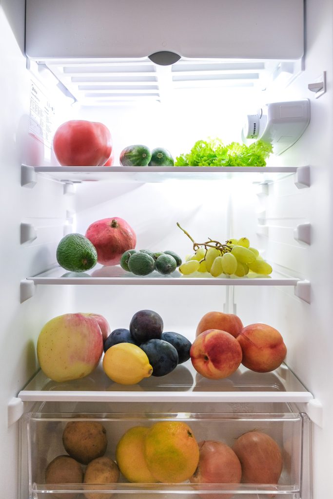 The lit interior of a tall refrigerator column, fruit and salad on glass shelves and a crisper drawer below