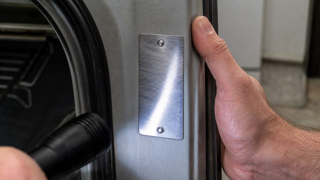 A blank stainless data plate riveted inside an appliance door frame, lit by a flashlight, a thumb holding the door open