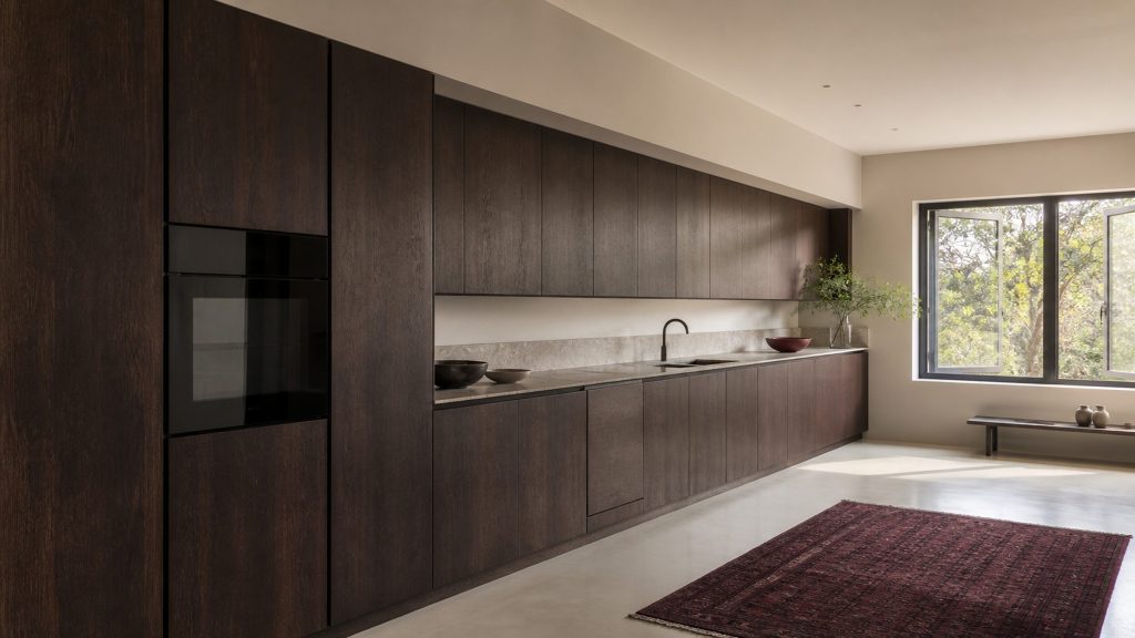 A long unbroken run of dark-stained oak kitchen cabinetry down one wall, integrated appliance doors flush in the column, a claret rug on the floor
