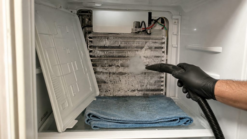 A gloved hand playing a steam nozzle over the iced evaporator coil behind the removed back panel of a freezer, a folded towel below