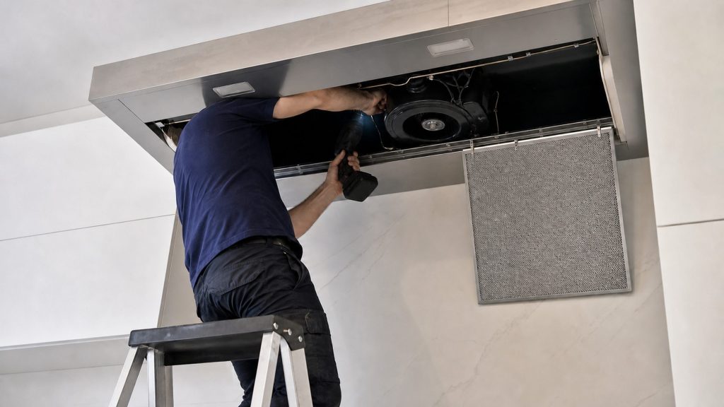 A technician on a step ladder reaching into the open canopy of a stainless range hood, one grease filter hanging down beside the blower