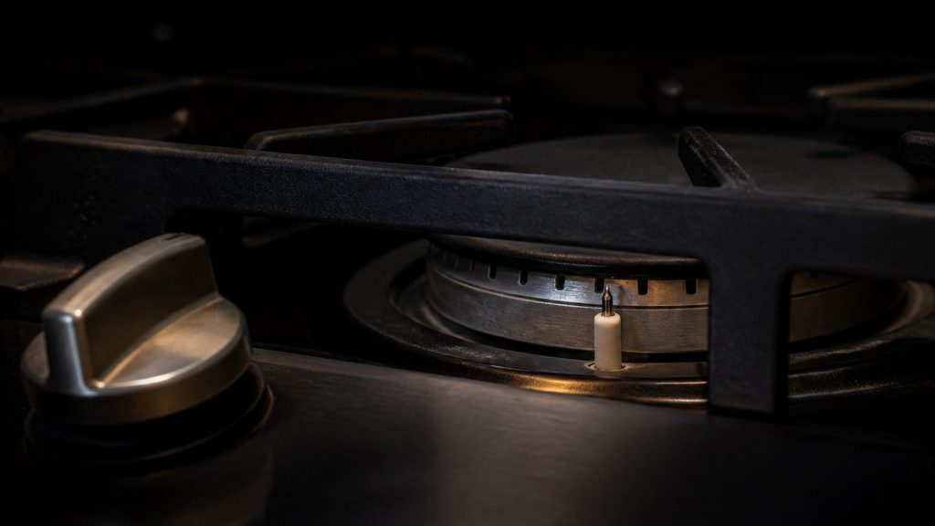 A gas burner ring and its white ceramic igniter under a cast grate, no flame lit, the knob turned on
