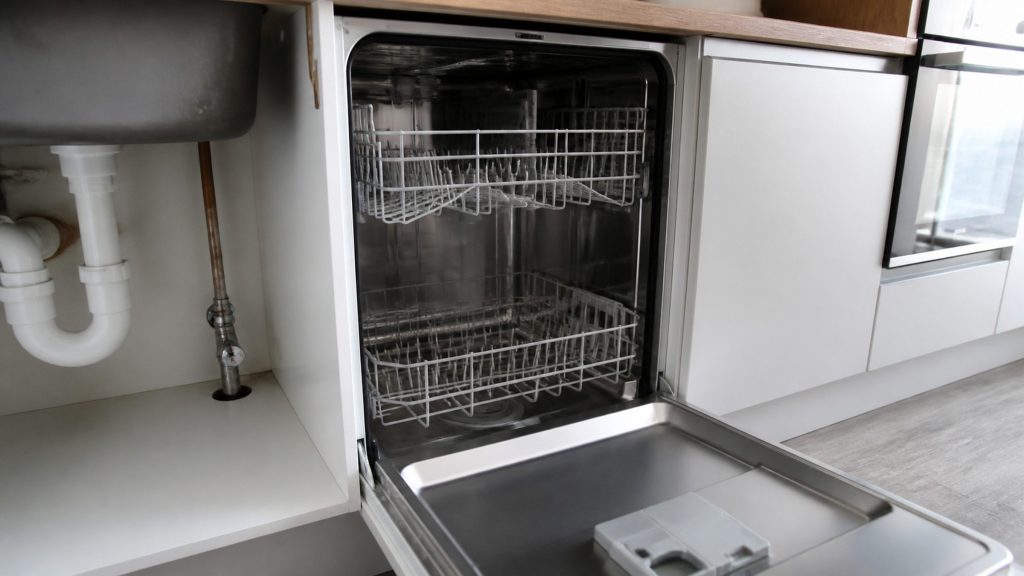 The empty dry tub of an integrated dishwasher standing open beside the under-sink cabinet, the water shutoff valve visible on the supply pipe