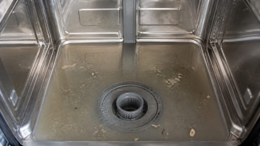 Cloudy water standing over the floor of a dishwasher tub around the filter, with food debris left in it